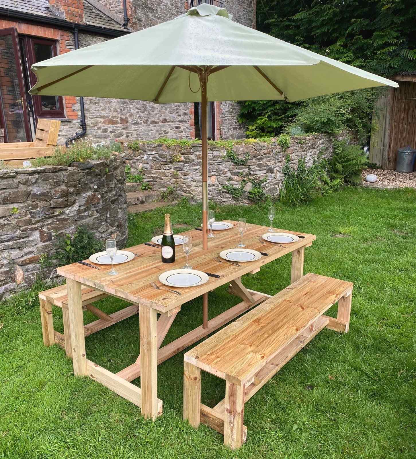 A heavy timber table and two benches set for dinner under a parasol on a lawn