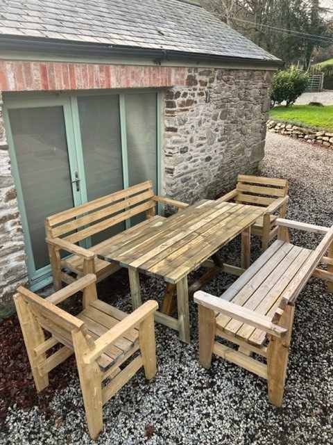 The Full Comfort 8 Seater set: a timber table with a bench and two armchairs on gravel by a stone barn