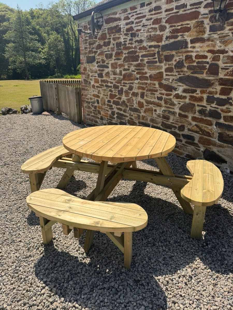 A round picnic table with curved benches on gravel by a stone barn